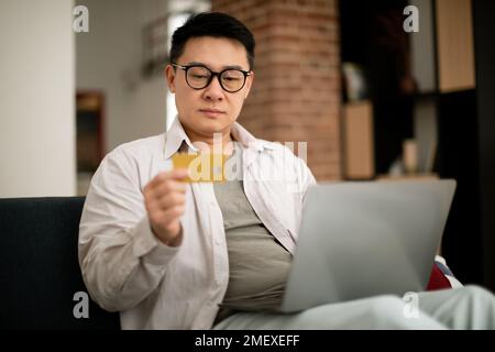 Portrait of asian mature man using pc and credit card, sitting on couch at home, free space Stock Photo