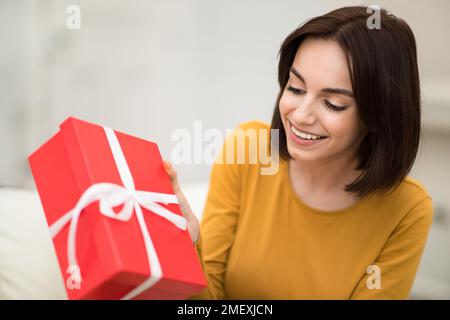 Cheerful young woman sitting on couch, holding cardboard gift box Stock Photo