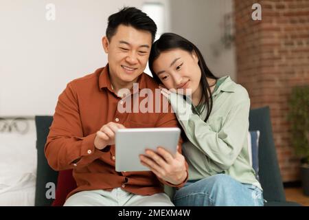 Happy asian spouses using digital tablet, watching videos or surfing internet, relaxing on couch in living room Stock Photo