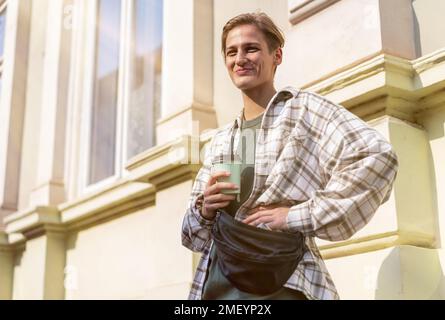 Young short blonde hair woman standing on street in city holding disposable coffee cup in hand looking at camera and smiling. Stock Photo