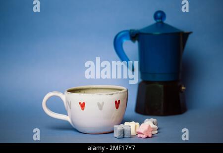 Small white cup in hearts with espresso coffee, cocoa or hot chocolate drink on blue background. Marshmallows on a table. Sweet dessert. Cozy still li Stock Photo