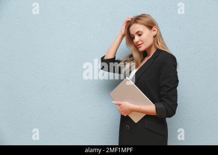 Blonde young business woman wearing black suit over blue plain background, carrying laptop Stock Photo