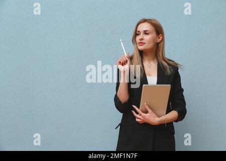 Blonde young business woman wearing black suit over blue plain background, carrying laptop Stock Photo