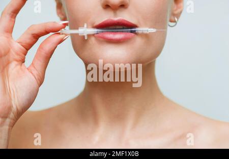Closeup face young woman holds syringe in mouth. Hyaluronic acid collagen injection for lips Stock Photo