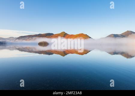 Mirror-like reflections of mountains in misty lake on a sunny blue sky morning. Derwentwater, Lake District, UK. Stock Photo
