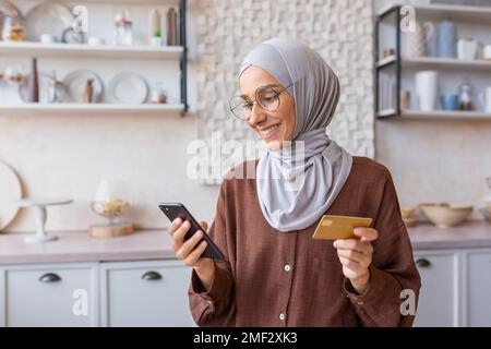 Beautiful muslim girl close up in kitchen, woman in hijab smiling and happy holding smartphone and gold bank credit card, shopping online in online store at home. Stock Photo