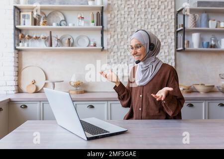Joyful woman in hijab working from home remotely, Muslim woman talking on video call using headset and laptop, businesswoman working remotely at home. Stock Photo
