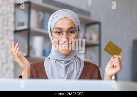 Cheerful and successful woman in hijab at home in kitchen with laptop and bank credit card makes online purchase in online store, Muslim woman chooses products and gifts remotely. Stock Photo