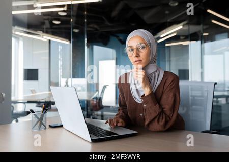 Portrait of a young woman Arab female student in hijab sitting in the office at the table and studying online at the laptop. He looks seriously at the camera. He holds his chin with his hand. Stock Photo