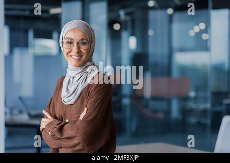 Portrait of young beautiful confident Arab business woman in hijab. Standing in the office, smiling, looking at the camera, crossed her arms. Stock Photo