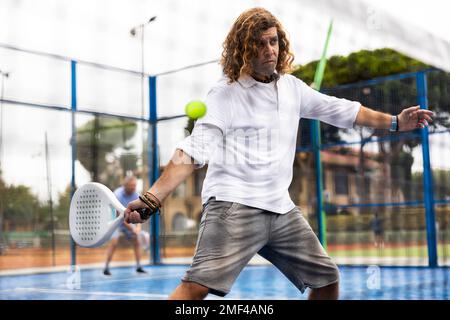 Focused middle-aged Latin man playing padel on court. View through tennis net Stock Photo