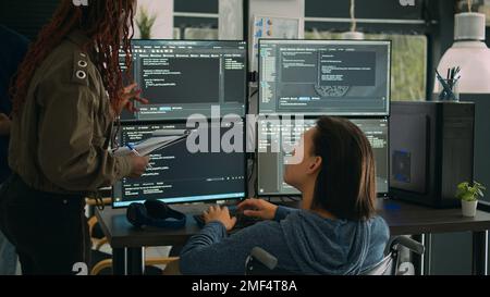 Female developer analyzing program code and talking to asian engineer with chronic disability, collaborating on new interface development. Wheelchair user and app developer programming language. Stock Photo