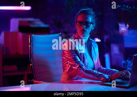 Neon metaverse futuristic concept. pensive trendy woman in glasses with laptop in modern office. Stock Photo