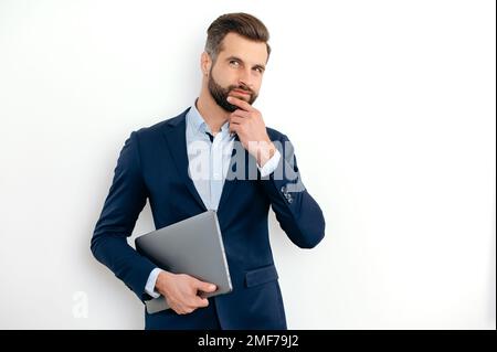 Successful confident caucasian businessman in a suit, seo, executive, broker, financial adviser, standing on isolated white background, holding laptop, looking to the side, thinking over idea,dreaming Stock Photo