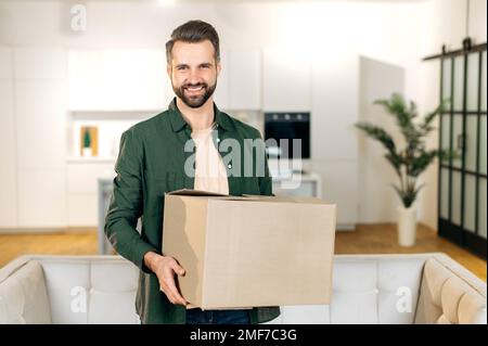 Happy attractive stylish caucasian man, stand at home in living room, holding in hands a large cardboard box, received parcel from the online store, preparing to unpack, looks at camera,smile friendly Stock Photo