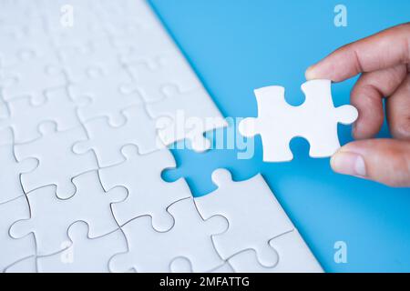 Hand put last piece of jigsaw puzzle. White unfinished jigsaw puzzle on  blue background.The correct solution. Teamwork, Solving and completing the ta Stock Photo