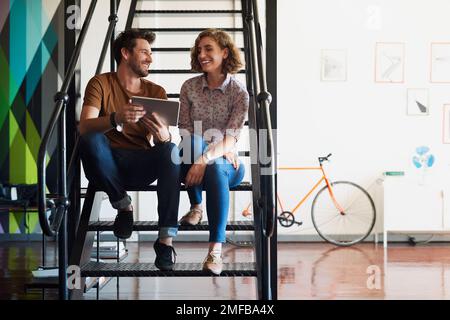 Using technology to bring their ideas together. two designers discussing something on a tablet while sitting on a staircase. Stock Photo