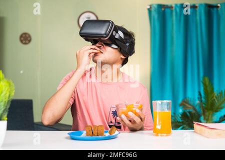 addicted Young man with VR or virtual reality having snacks on metaverse while sitting at home - concept of futuristic, modern lifestyle and Stock Photo