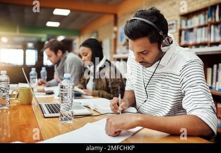 Hes focused on his studies. a young male university student studying in the library with other students in the background. Stock Photo