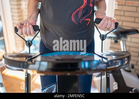 Older white woman doing exercises with her arms on a machine that vibrates to keep fit at home. Stock Photo