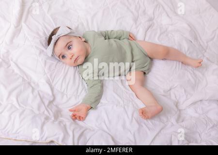 a girl with beautiful big eyes is rubbing the baby at home on the bed in a cotton bodysuit on white bed linen. High quality photo Stock Photo
