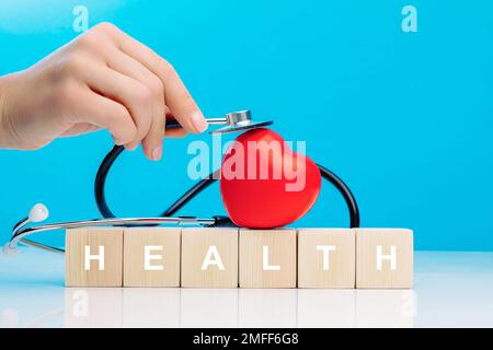A woman's hand holds a stethoscope and touches a red heart that lies on top of wooden cubes with the inscription HEALTH Stock Photo