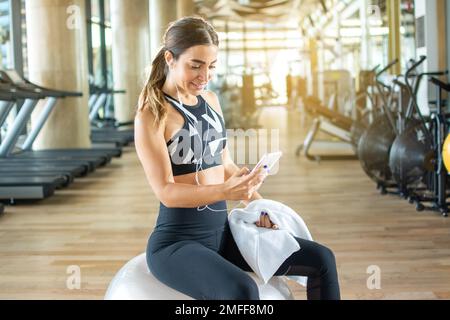 Beautiful woman in sportswear wearing headphones listening to music on smartphone after sports training at gym. Stock Photo