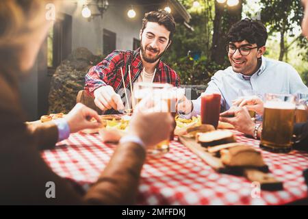 A group of friends gather around a checkered table in a backyard patio, laughing and enjoying beers and nachos on a sunny day Stock Photo
