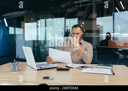 Thoughtful young African-American male designer, architect sitting in the office at the table with a laptop. He holds documents in his hands, concentrates on working with plans and drawings. Stock Photo