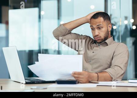 Worried young African American male student holding papers in hands. I received a letter, bad exam results, refusal of admission. Upset, holding his head. Stock Photo
