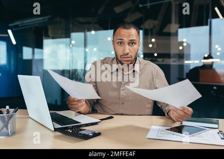 Problem with documents at work. Upset young African-American businessman working in office, holding papers in hands, spreading hands, looking worriedly at camera. Stock Photo