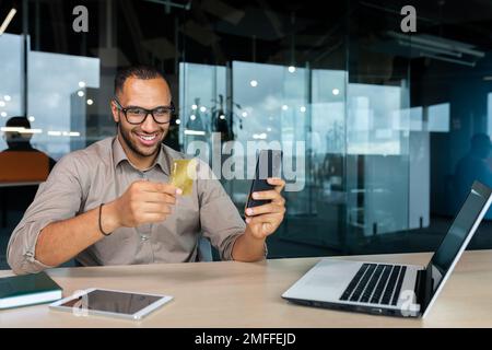 Smiling young African American man sitting in office at desk with laptop. He is holding a credit card, using the phone. Stock Photo