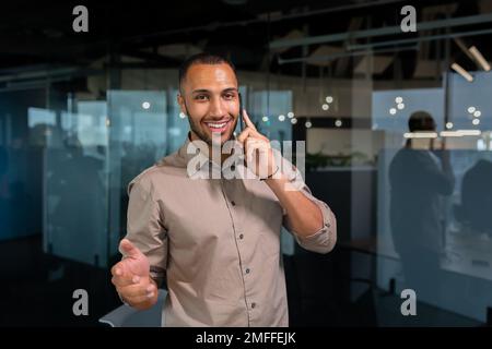 A young African American man is standing in the office and talking on the phone. gesturing with his hands, looking at the camera, smiling. Stock Photo