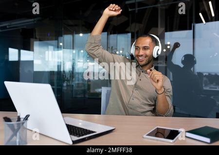 Happy young African American man sitting in office at desk with laptop wearing white headphones. He listens to music, dances, raised his hands up, rests, a break. Stock Photo