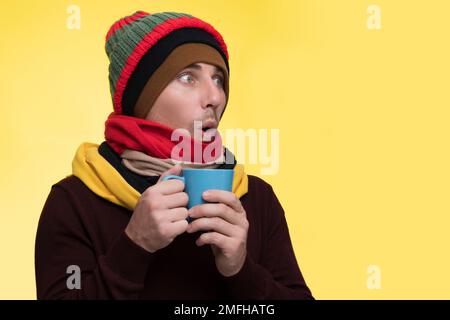 A man on a yellow background in several hats and scarves holds a cup in his hands, cold winter and warm outerwear. Stock Photo