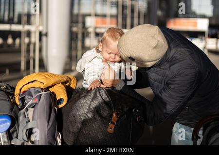 Fatherat comforting his crying infant baby boy child tired sitting on top of luggage cart in front of airport terminal station while traveling wih family Stock Photo