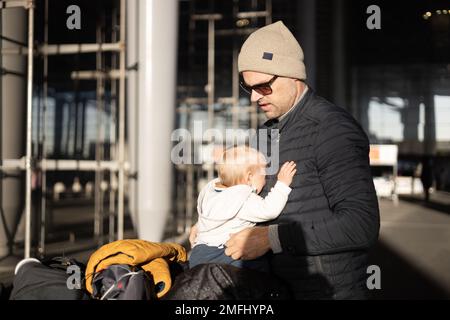 Fatherat comforting his crying infant baby boy child tired sitting on top of luggage cart in front of airport terminal station while traveling wih family Stock Photo