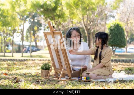 Happy and lovely Asian grandmother teaching her granddaughter to paint watercolors on canvas, having a great time together in the beautiful park Stock Photo