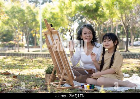 Happy and lovely Asian grandmother teaching her granddaughter to paint watercolors on canvas, having a great time together in the beautiful park Stock Photo