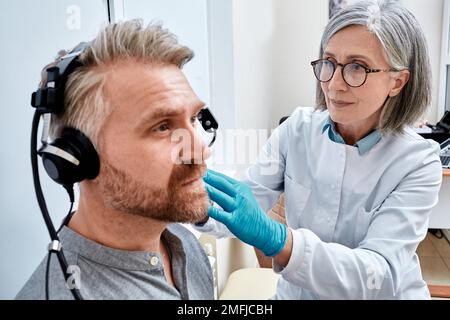 Mature man receives tympanometry with audiologist using tympanometer at audiology clinic. Hearing check-up, impedance audiometry Stock Photo