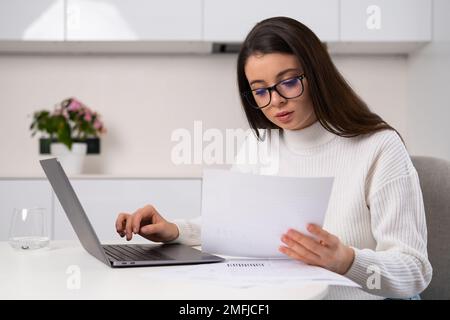 Focused woman enters data written in papers into Notebook sitting at table in modern kitchen. Young female freelancer in glasses enjoys working online Stock Photo