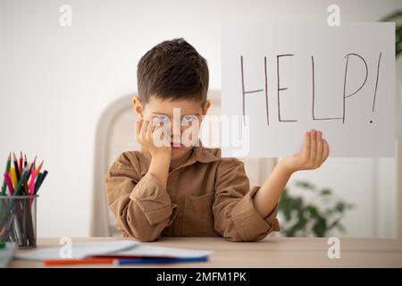 Sad unhappy little boy sits at table, studies and shows banner with inscription help in room interior, free space Stock Photo