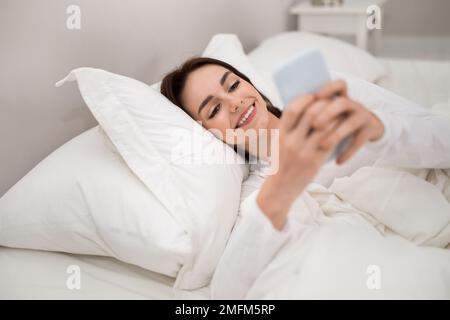 Cheerful young woman using phone in bed Stock Photo