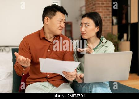 Angry korean spouses quarreling over debts and bills with papers and laptop, sitting on sofa at home Stock Photo