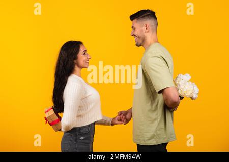 Smiling young arab guy hold bouquet behind his back and woman with gift box celebrating holiday Stock Photo