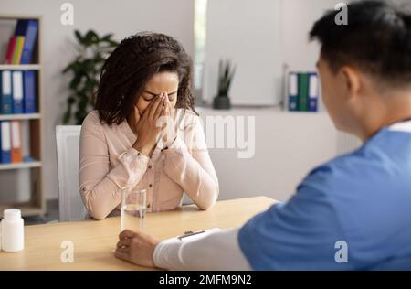 Serious middle aged korean doctor calms crying black millennial female patient, giving glass of water Stock Photo