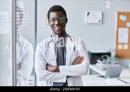 Waist up portrait of confident black doctor leaning on glass wall in office and smiling at camera, copy space Stock Photo