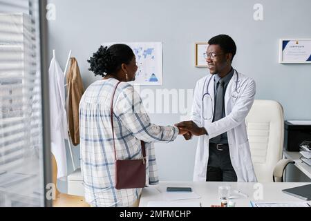 Portrait of smiling black doctor holding hands with female patient during friendly greeting for consultation in private clinic Stock Photo