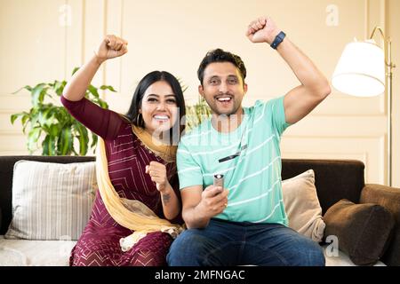 Excited cheerful couple shouting by rising hands while watching live cricket sports match on tv or television at home - concept of entertainment Stock Photo