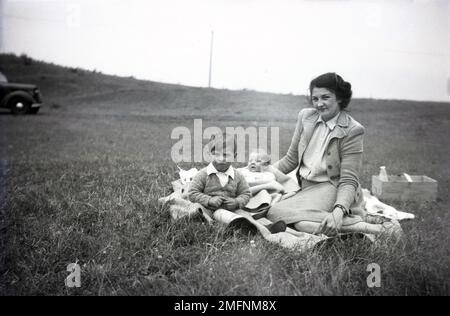 1950s, historical, a mother sitting on a blanket in a field, with her young two sons, a small boy and infant child, England, UK. Beisde her, a milk bottle in wooden box. The family car of the era is seen on the left. Stock Photo